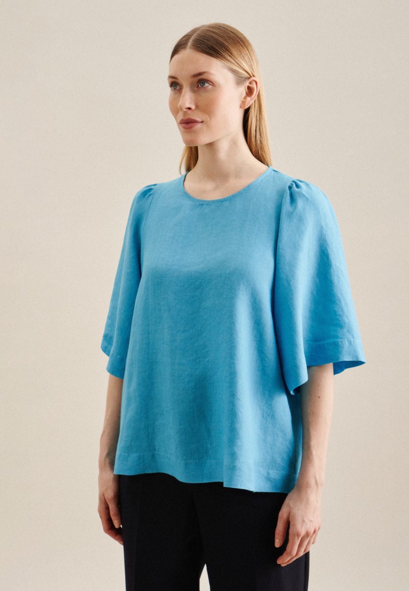 Top in lino azzurro chiaro con maniche corte e puffate e scollatura rotonda; vestibilità morbida con una texture liscia e dettagli di cucitura minimi.