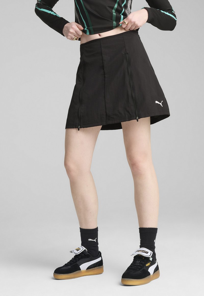Jupe de sport noire avec deux zips latéraux, fabriquée en matériau léger, associée à des baskets noires et blanches avec un logo.