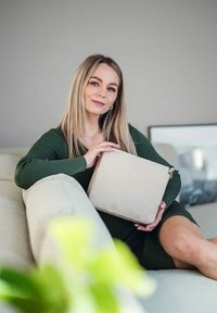 Beige, texturovaná kabelka se zipovým uzavřením, držena na klíně ženy, na neutrálním pozadí. Viditelný zelený svetr a měkká pohovka.