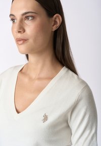 Maglione bianco a V in materiale morbido, con un piccolo logo ricamato sul lato sinistro del petto. Texture liscia e design aderente.