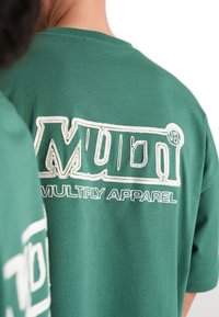 T-shirt verde a maniche corte con il testo ricamato in bianco "MULTI" e "MULTIPLY APPAREL" sul retro. Tessuto morbido, vestibilità rilassata.