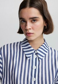 Camisa de cuello con rayas verticales en azul marino y blanco, con botones en el frente y una textura de algodón suave. Vista en primer plano enfocada en el cuello.