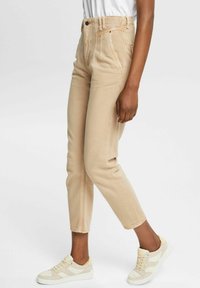 Pantalons beiges taille haute en coupe ajustée, avec poches avant et détails cousus, associés à des baskets de sport de couleur claire.