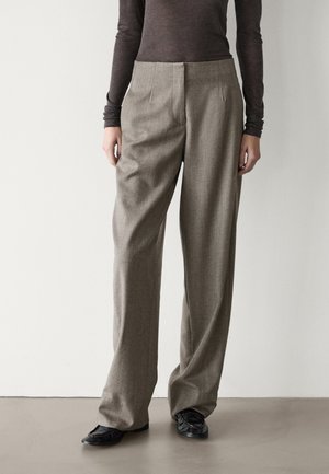 Personne portant un pantalon beige à taille haute et jambes larges, un haut marron à manches longues et des chaussures plates noires, debout devant un mur blanc uni.