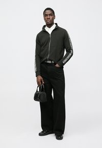 Black zip-up jacket with white side stripes, paired with wide black pants. Hold a small black bag. White collared shirt underneath.