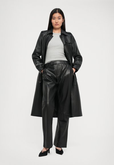 Woman stands with hands in pockets wearing black leather trench coat, black leather trousers, gray ribbed top, and black pointed heels.