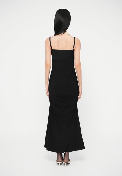 Sieviete ar gariem melniem matiem, tērpusies pieguļošā melnā vakarkleitā ar plānām lencītēm, valkā pērļu krelles, melnas zeķubikses un baltus augstpapēžu apavus, stāvot ar muguru.