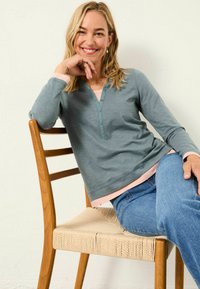 Langärmliges Oberteil aus weichem Tön in Teal, mit einem Henley-Ausschnitt und Knöpfen, über einem hellrosa Shirt getragen, kombiniert mit blauen Jeans.