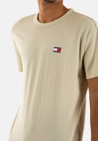 Maglietta beige a maniche corte in cotone, con una piccola toppa logo rettangolare blu, bianca e rossa sul lato sinistro del petto. Collo rotondo.