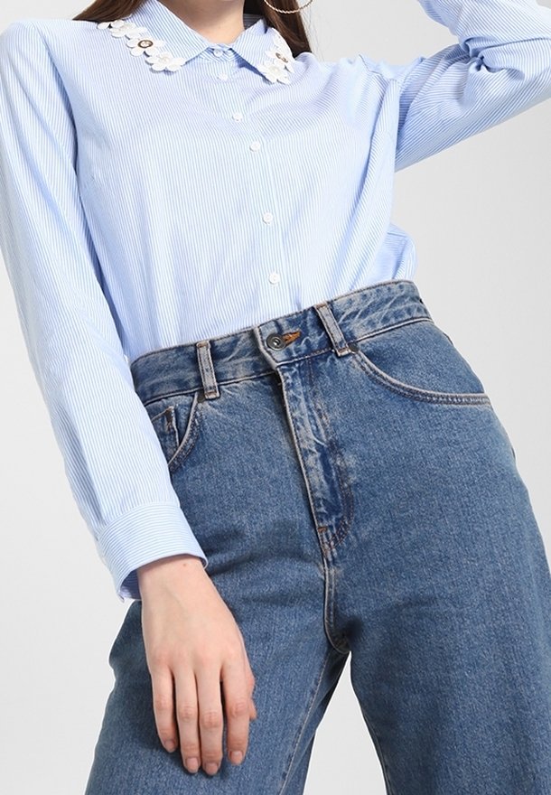 Vaaleansininen raidallinen paita, jossa on valkoinen koristeellinen kaulus, yhdistetty korkeavyötäröisiin sinisiin farkkuihin, joissa on nappikiinnitys ja etutaskut.