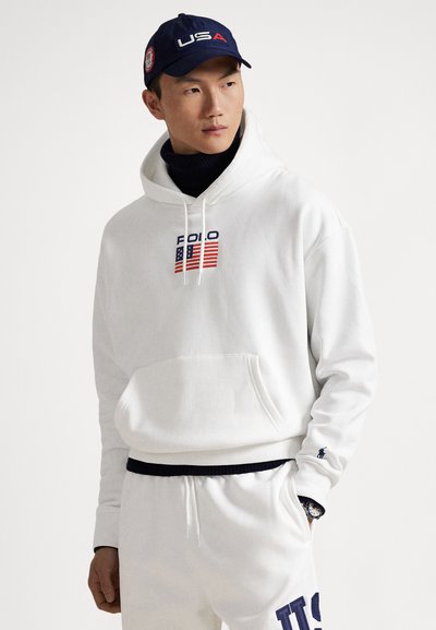 White hooded sweatshirt with red and blue "POLO" text and American flag design. Paired with white pants and a blue baseball cap featuring "USA."
