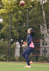 Sportaš u tamnoplavoj i ružičastoj sportskoj odjeći, žonglira crno-crvenom nogometnom loptom na zelenom terenu s lančastom ogradom u pozadini.