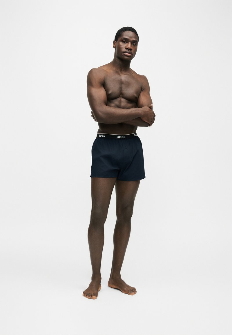 Homme musclé debout pieds nus, bras croisés, portant un boxer bleu foncé avec une ceinture de marque.