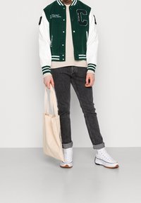 Giacca varsity verde e bianca con toppa a forma di "C", jeans neri, sneakers bianche con dettagli marroni e una borsa a tracolla beige.
