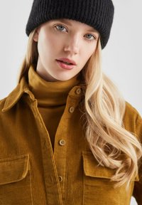 Senfgelben Corduroy-Hemd mit Knopfleiste und zwei Brusttaschen, getragen über einem gelben Rollkragenpullover, kombiniert mit einer schwarzen gerippten Strickmütze.