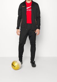 Osoba v černém sportovním oblečení a červeném tričku stojí s jednou nohou vedle zlatého a bílého fotbalového míče na čistém bílém pozadí.