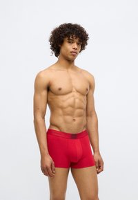 Boxers ajustados de algodão vermelhos com cintura elástica destacando o logótipo. Design ajustado, textura suave e comprimento de perna curto. Modelo de pé com os braços relaxados.