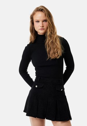 Młoda kobieta z długimi blond włosami, ubrana w czarny prążkowany golf i plisowaną czarną spódnicę z patkami kieszeni.