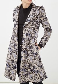 Abrigo con estampado floral que presenta un fondo beige claro con diseños en negro y azul. Destaca por su cuello con puntas y bolsillos laterales.