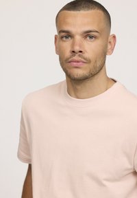 T-shirt en coton rose pâle à col rond, à manches courtes, présentant une texture lisse et un design minimaliste. Pas de logos ou de motifs visibles.