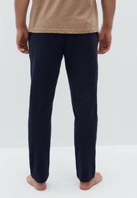Marineblauwe broek van zachte stof, met een recht gesneden ontwerp en een enkele achterzak. G gedragen met een beige shirt en blote voeten.