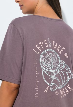 Vrouw draagt een mauvekleurig shirt met afbeelding van een koffiekopje, croissant en de tekst 'Let's take a break' op de achterkant.