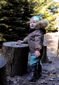 Enfant portant une parka marron à capuche bordée de fourrure, portant un pantalon en polaire turquoise et des bottes en caoutchouc noires, debout près d'une souche en bois dans une forêt.