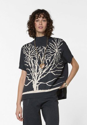 Frau mit lockigem Haar, die einen dunkelblauen Kurzarm-Pullover mit einem großen weißen Baum-Muster, dunkle Hose, Herz-Halskette und Armbanduhr trägt.