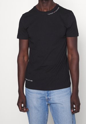 Mand iført sort Tommy Hilfiger T-shirt med logo nær kraven og lyseblå jeans, stående mod en ensfarvet baggrund.
