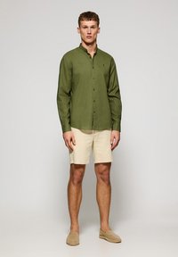 Groene knoopshirt met een kraag, beige shorts en lichtbruine espadrilles, staand tegen een neutrale achtergrond.