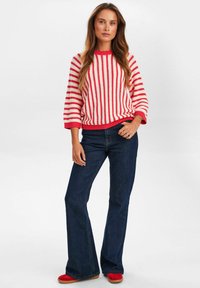 Czerwono-biały sweter w paski z szerokimi rękawami, w zestawieniu z ciemnoniebieskimi dżinsami z rozszerzanymi nogawkami i czerwonym obuwiem. Dzianina o teksturze z prążkowanymi akcentami.