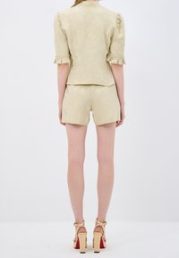 Femme portant un haut beige texturé à manches courtes avec des poignets à volants et un short assorti, accompagné de sandales beiges à talons hauts avec des brides à la cheville.