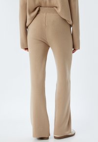 Pantaloni beige a zampa d'elefante in un tessuto morbido e testurizzato, con un cinturino a coste e una finitura liscia, abbinati a scarpe chiare.