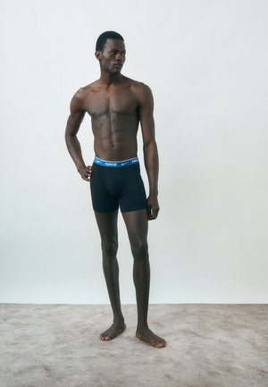 Άνδρας μαύρος χωρίς πουκάμισο, γυμνό πόδι σε δαπέδο με μοκέτα, φοράει μαύρα boxer briefs Nike με μπλε ζώνη, κοιτάζει προς τα πλάγια.