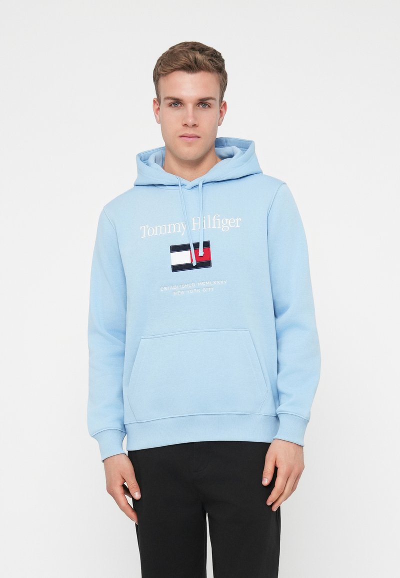 Απαλή γαλάζια φόρμα με κουκούλα και εμπρόσθια τσέπη τύπου καγκουρό, με σχέδιο λογότυπου και σημαίας στο στήθος. Κατασκευασμένη από μαλακό ύφασμα μίξης βαμβακιού.