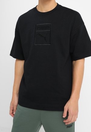 Homme portant un t-shirt noir Puma avec logo embossé et un pantalon de survêtement Puma vert olive, debout devant un fond uni.