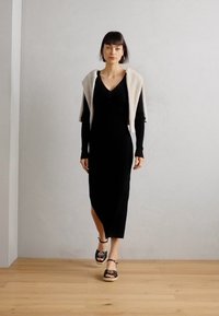 Svart ribbad stickad klänning med längd till mitten av vaden och en sidslits, kombinerad med en ljusgrå draperingströja och svarta sandaler med remmar.