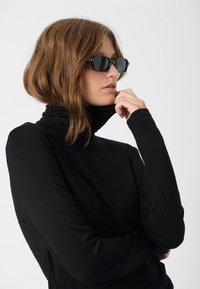 Black turtleneck top with long sleeves, made from a smooth, stretchy fabric. Model wears dark rectangular sunglasses. Simple, fitted design.