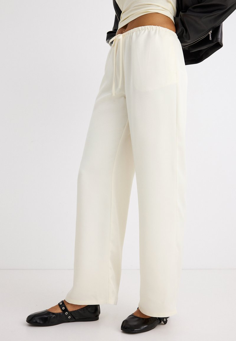 Γυναίκα που φοράει άνετο κρεμ παντελόνι με κορδόνι και μαύρα επίπεδα παπούτσια, στέκεται μπροστά σε ένα απλό λευκό φόντο.