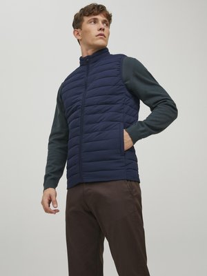 Marineblaue gesteppten Weste mit hohem Kragen, Reißverschluss vorne und seitlichen Taschen, über einem dunkelgrünen Langarmshirt und braunen Hosen getragen.