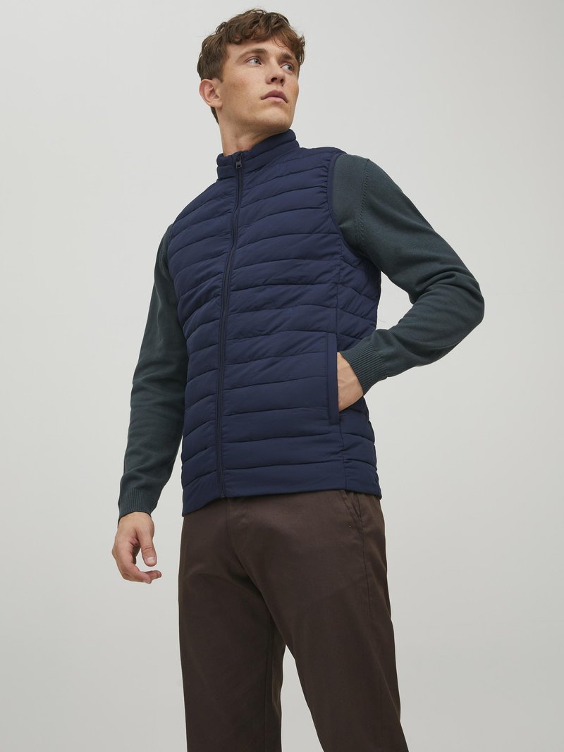 Gilet matelassé bleu marine avec col haut, fermeture éclair à l'avant et poches latérales, superposé à une chemise à manches longues vert foncé et un pantalon marron.