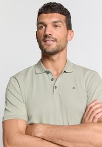 Lichtgrijze poloshirt met een kraag, drie donkere knopen, korte mouwen en een subtiel logo op de borst. Gladde stoftextuur.