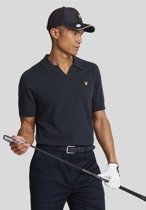 Polo noir à manches courtes avec un col en V et un petit logo jaune, associé à un pantalon marine et une casquette noire, tenant un club de golf.