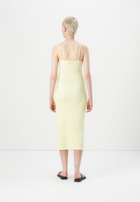Robe midi jaune à côtes avec des bretelles fines et une silhouette ajustée, dotée d'un détail de bouton dans le dos, portée avec des sandales plates noires.