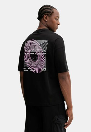 Uomo con capelli intrecciati che indossa una maglietta nera con design geometrico rosa e bianco e testo sul retro, in piedi davanti a uno sfondo bianco.