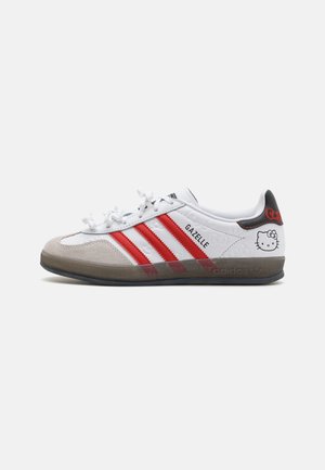 Biały sneaker Adidas Gazelle z czerwonymi paskami, szarą zamszową czubką, czarną zakładką na pięcie i detalem twarzy Hello Kitty po boku.