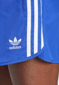 Modré sportovní šortky vyrobené z lehkého materiálu, s třemi bílými pruhy po straně a vyšitým bílým logem Adidas.