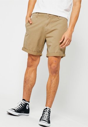 Mann trägt hochgekrempelte beige Shorts, weißes T-Shirt und schwarze High-Top-Sneakers, steht vor weißem Hintergrund.