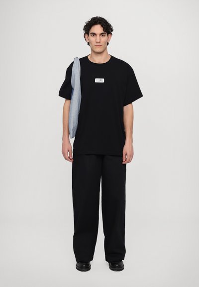 T-shirt noir surdimensionné avec un patch graphique blanc, associé à un pantalon noir ample. Le mannequin tient un pull bleu clair drapé sur une épaule.