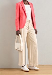 Blazer corail, top blanc ajusté, pantalon beige à jambes larges et baskets blanches ; tenant un sac crème avec un rabat et des détails en métal argenté.